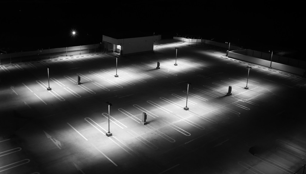 Black-and-white photograph by Mitsutaka Tsuryu showing an empty parking lot at night, viewed from above, illuminated by evenly spaced streetlights.